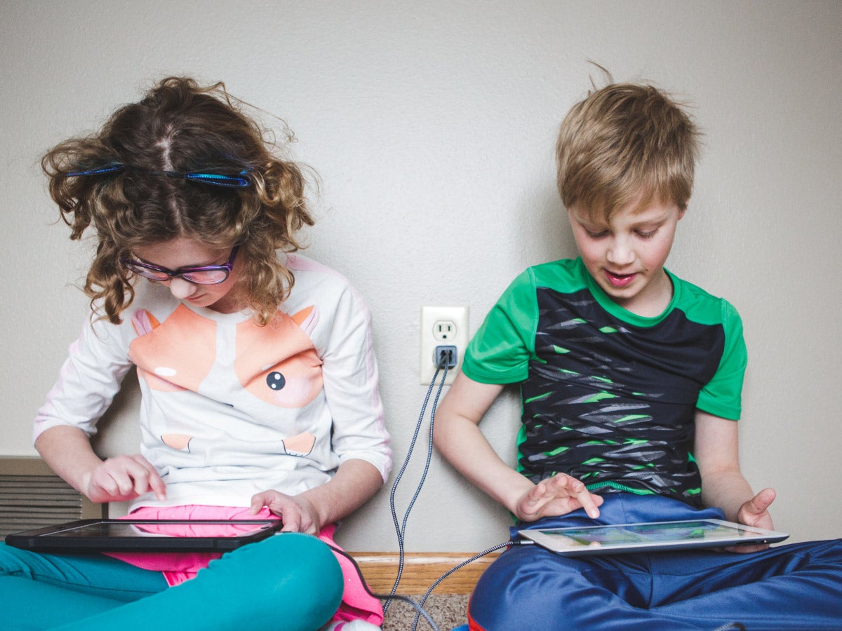 Two children sit side by side against a wall and look down at their digital tablets that are both plugged into a the same wall charger.